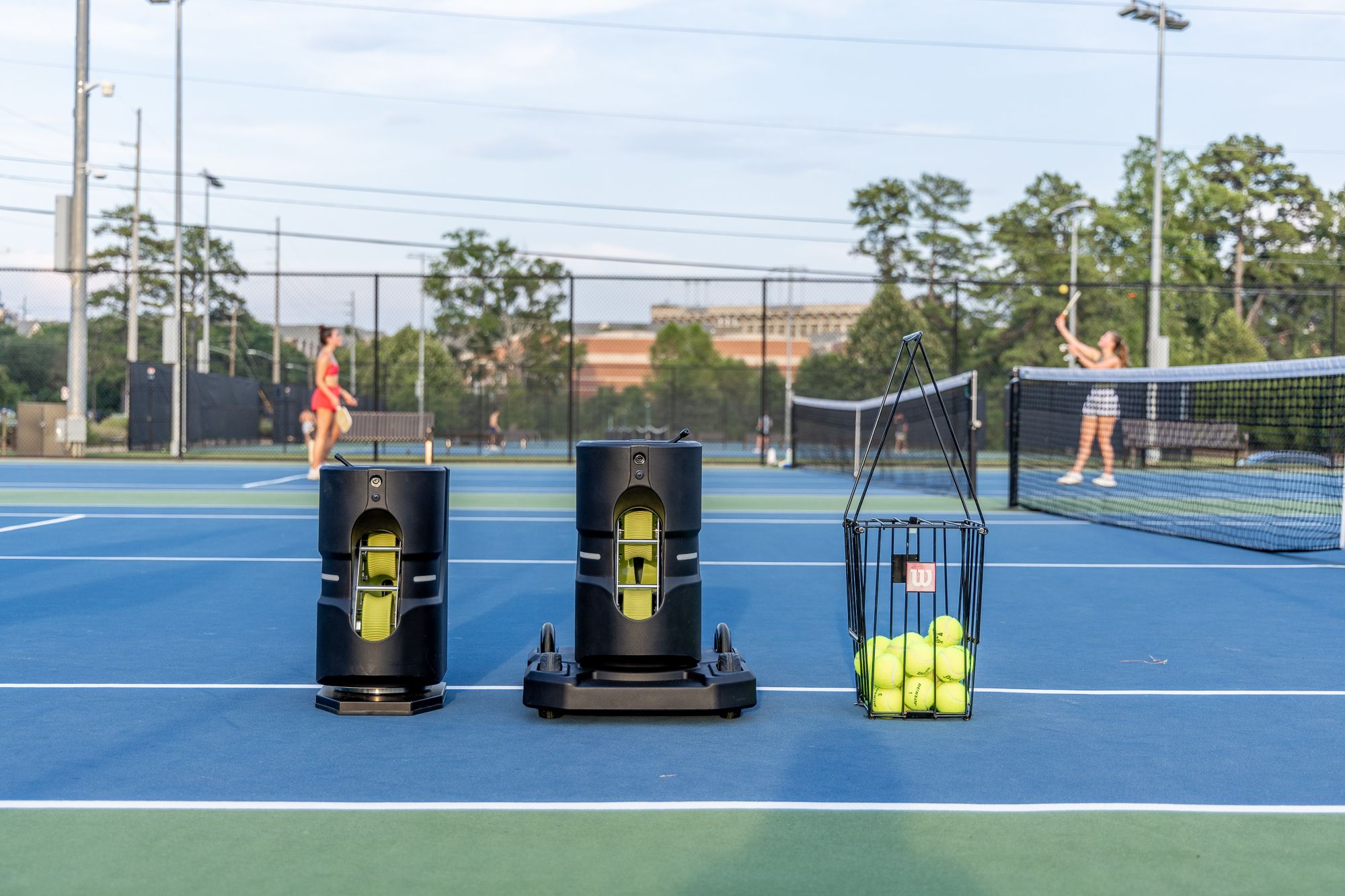 Tennibot Partner Lite and Partner V2 ball machines next to a basket of tennis balls on a hard court