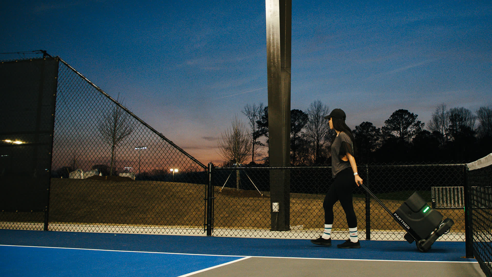 A player pulling the Tennibot Partner V2 by its handle across a tennis court at dusk