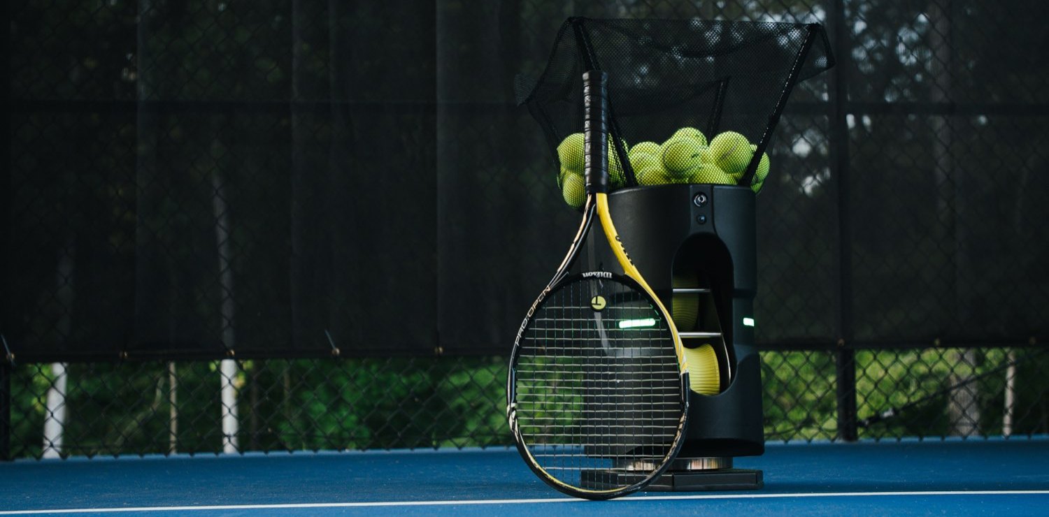 An empty tennis court at dusk with a racquet and a basket of balls waiting at the baseline